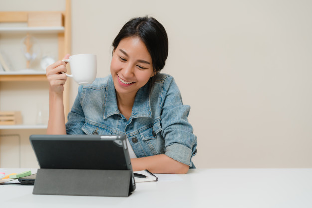 young-asian-woman-working-using-tablet-checking-social-media-drinking-coffee-while-relax-desk-living-room-home_7861-1385